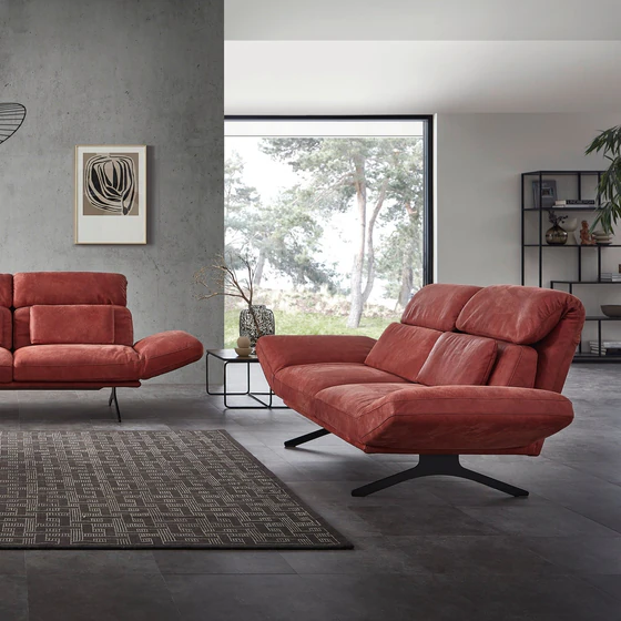 Relaxsofa Meda von Nolte Polster in Amarena-Rot mit individuell verstellbaren Armlehnen – modernes Design in offenem, lichtdurchflutetem Wohnraum mit Betonwand und großen Fensterfronten.