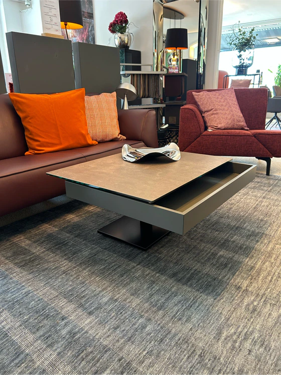 Stylischer Funktions-Couchtisch Ronald Schmitt Janas K 471 mit Duo‑Höhenverstellung, aufgeschobene Lade rollbar und verschiebbarer Lade im Einrichtungshaus Zeppenfeld in Olpe.