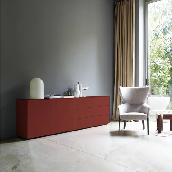 Sideboard in kräftigem English Red in einer modernen Wohnumgebung mit grauer Wand, hellem Boden und bequemen Sessel am Fenster.