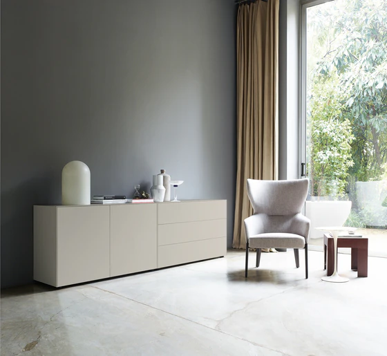 Wohnsituation mit Nex Pur Box Sideboard in komplettem Seidengrau: links Türmodul 1536778, rechts Schubkastenelement 1536776, vor grauer Wand mit großer Fensterfläche und Designstuhl.
